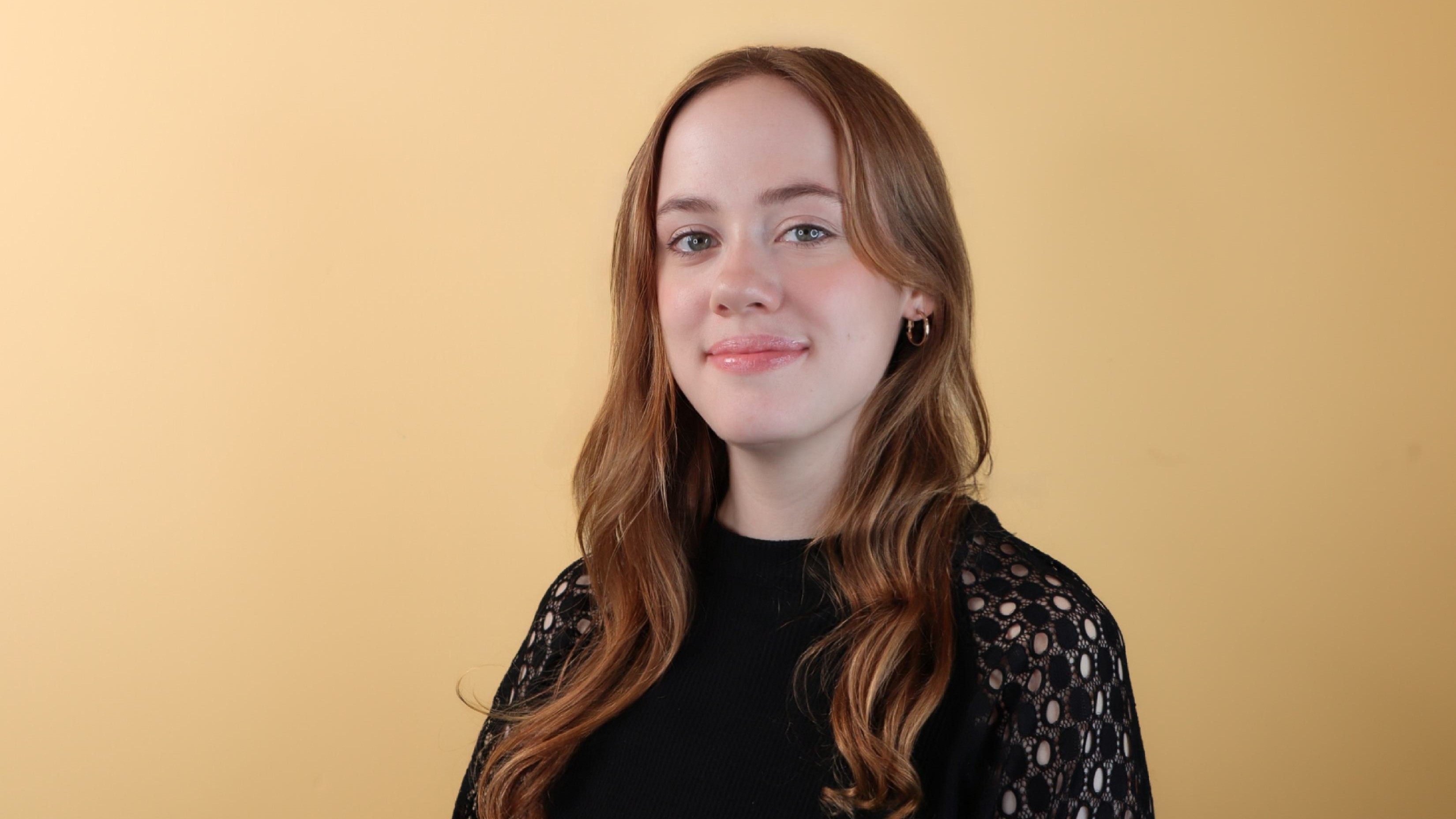 Zoe Schmidt smiles in front of a beige background while wearing a black long sleeved shirt. Zoe Schmidt smiles in front of a beige background while wearing a black long sleeved shirt.